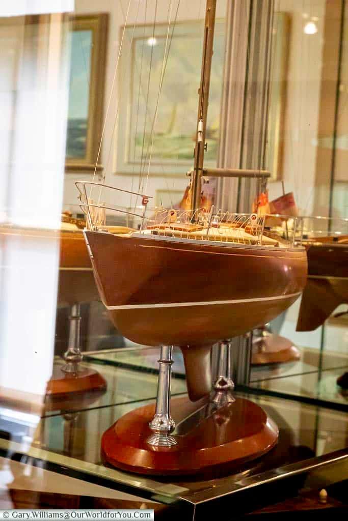 Captured through a glass display case at Arundells House in the UK, a meticulously crafted model sailboat with a rich wooden hull, white accents, and delicate rigging is showcased on a polished wooden base with silver supports. Reflections on the glass hint at the elegant surroundings of the exhibition.