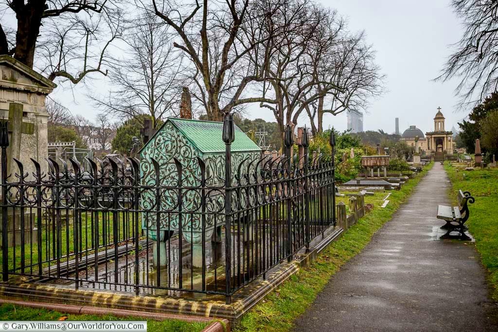 This wide shot shows a pathway winding through Brompton Cemetery in the United Kingdom on an overcast day, lined with various aged gravestones and monuments, some enclosed by ornate iron railings. In the background, bare trees frame a distant view of city buildings and the cemetery's chapel dome.