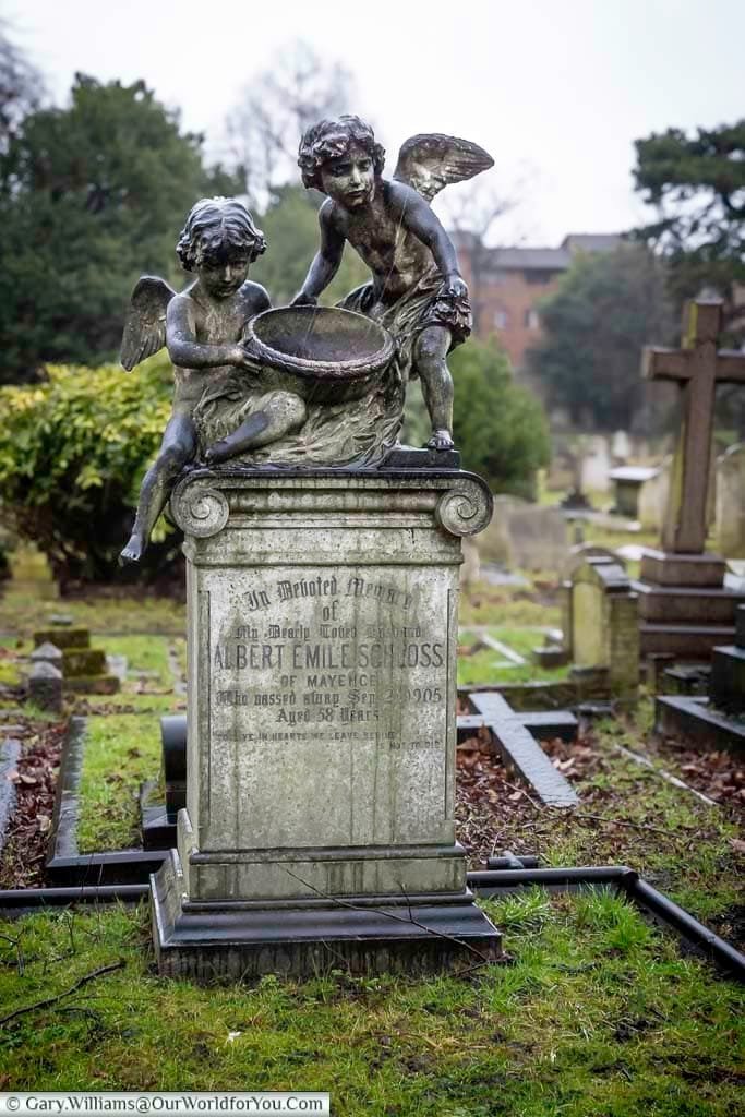 This close-up shot features a weathered stone monument in Brompton Cemetery, United Kingdom, topped with a bronze sculpture of two winged cherubs holding a basin. The pedestal bears an inscription "In Beloved Memory of ALBERT EMILE SCHLOSS," detailing his birth and death dates.