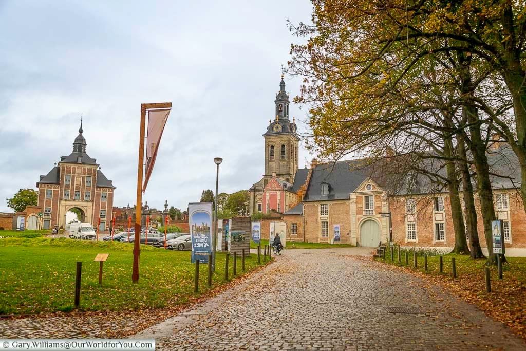 This eye-level, outdoor shot captures a cobblestone path leading towards the historic Park Abbey in Leuven, Flanders, Belgium, under an overcast sky. The abbey complex includes buildings with brick and stone facades, a prominent tower, and is surrounded by autumn foliage and parked cars.
