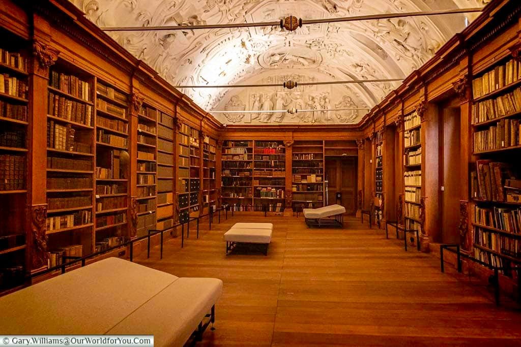 This is an interior view of the historic library at PARCUM, a museum in Leuven, Flanders, Belgium, showcasing religious art and culture. The long, narrow room features tall wooden bookshelves lining both sides, filled with old books, and several light-colored benches for visitors to sit and explore the collections under the ornate, vaulted ceiling.