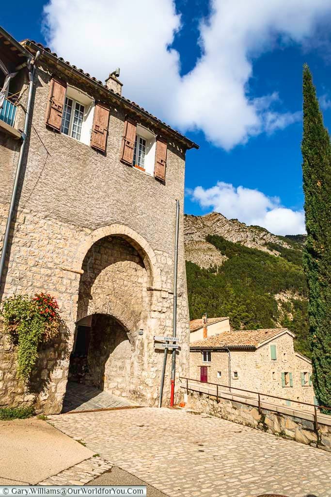 A charming view of Place de la Nière in Sisteron, Provence, France, showcases a historic stone building with a prominent archway and traditional wooden shutters, set against a backdrop of a tree-covered mountain and a brilliant blue sky with fluffy clouds. The sloping cobblestone ground, a tall, slender cypress tree, and other rustic buildings in this hidden gem contribute to the authentic French village atmosphere, perfect for a scenic road trip.