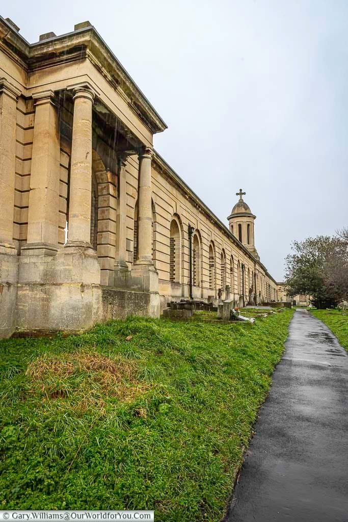 This image shows a long, colonnaded building with classical architectural elements in Brompton Cemetery, United Kingdom, under an overcast sky. A paved pathway runs alongside a grassy area with scattered gravestones, leading towards a domed structure with a cross in the distance.