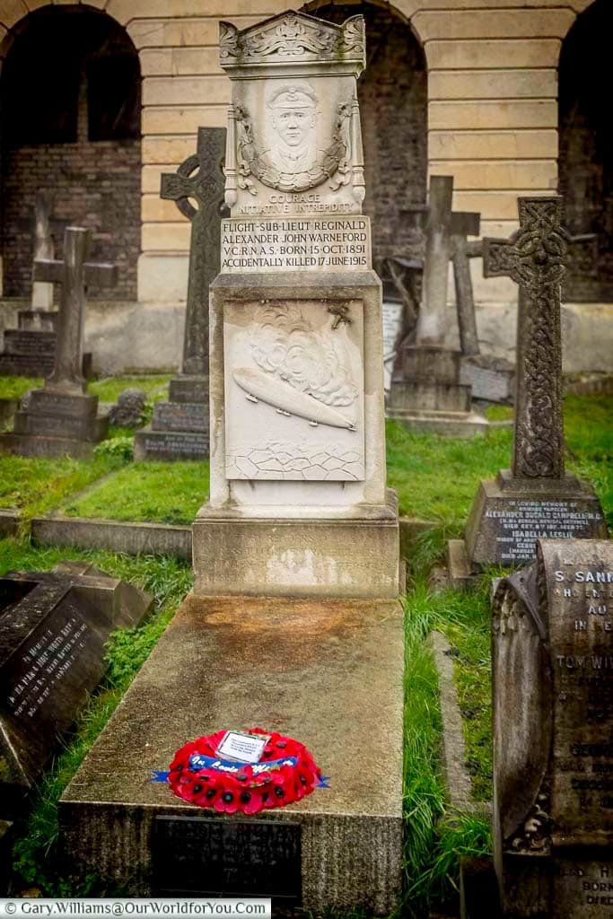 This image shows the gravestone of Flight-Sub-Lieutenant Reginald Alexander John Warneford VC in Brompton Cemetery, United Kingdom. The stone features a portrait, inscriptions detailing his service and death in 1915, and a relief carving of an airship being attacked, with a wreath of poppies and blue flowers laid at its base.