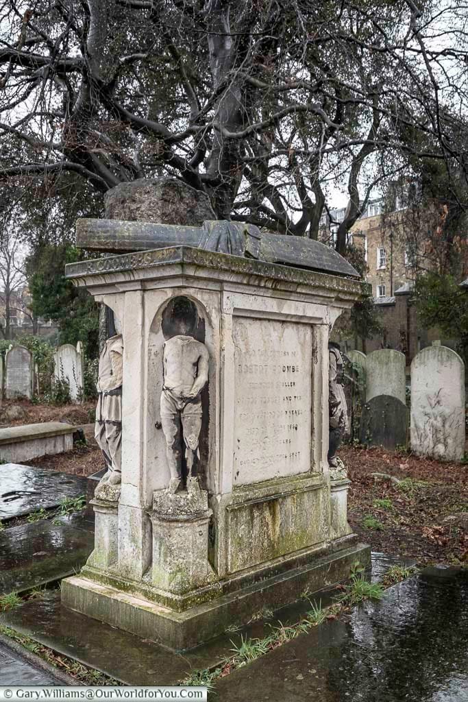 This image shows a weathered, multi-sided stone monument in Brompton Cemetery, United Kingdom, featuring carved figures in niches on each side and inscriptions detailing the deceased. The monument stands on a wet ground, surrounded by other gravestones and bare trees under an overcast sky.