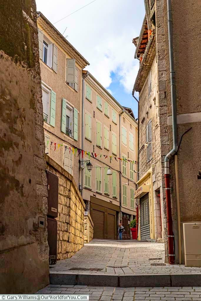 A charming, narrow cobblestone street, Rue du Glissoir in Sisteron, Provence, France, winds uphill between tall, historic buildings adorned with light-green shutters and festive bunting strung overhead. This picturesque French alleyway, with its textured stone walls and glimpse of blue sky, offers a quintessential view of hidden gems to discover while touring France.