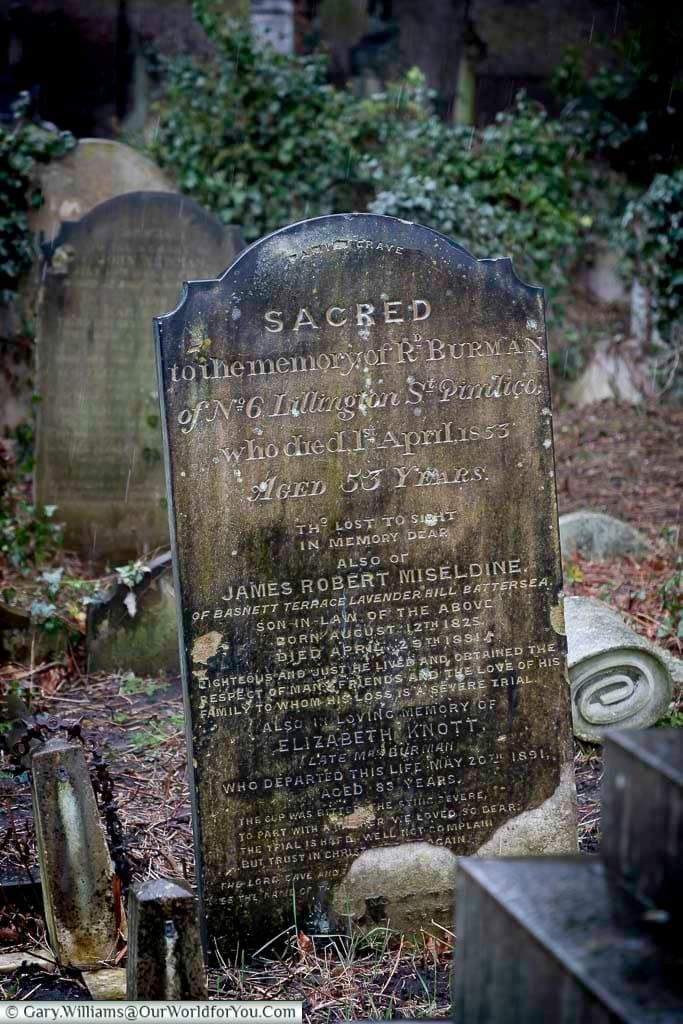 This image shows a weathered, upright gravestone in Brompton Cemetery, UK, surrounded by other graves and greenery. The inscription on the stone is partially visible, indicating it is a memorial.