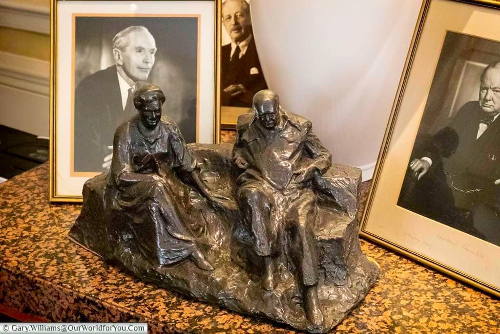 A bronze sculpture depicting two seated figures, winston churchill & his wife clementine, is displayed on a speckled brown surface at Arundells House in Salisbury. The sculpture is flanked by framed black and white portraits of distinguished-looking men, adding to the historical ambiance of the setting.