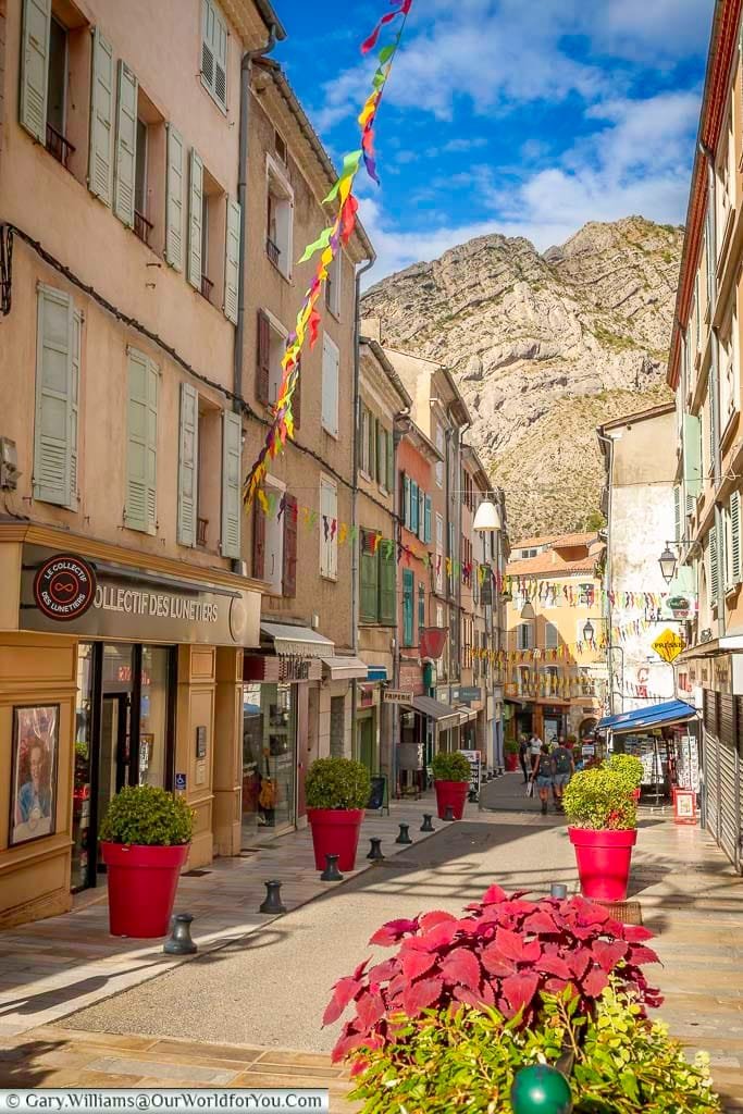 A charming, narrow street in Sisteron, France is lined with colorful buildings, local shops, and festive bunting strung overhead. The vibrant scene captures the lively atmosphere of this Provençal town under a bright, partly cloudy sky.