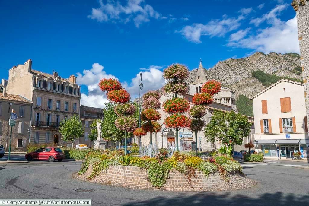 A vibrant roundabout in Sisteron, France, features a stunning floral display with trees topped by large globes of red and pink flowers, all set against a backdrop of historic town buildings and a distant rocky mountain. The scene is captured under a bright blue sky with scattered white clouds, with a street and parked cars visible around the roundabout.