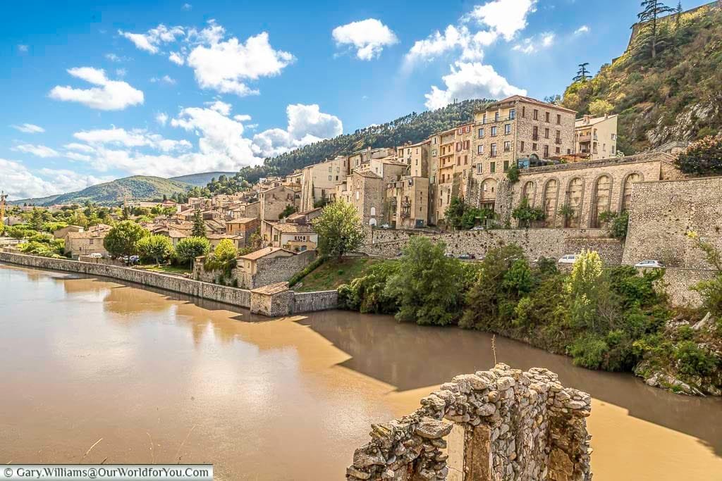 A picturesque view captures Sisteron’s medieval old town in Provence, France, with its historic buildings tightly packed along the hillside overlooking the wide, flowing Durance River. In the foreground, the stone ruins of an old structure add to the historic charm, all set against a backdrop of rolling hills and a bright blue sky with scattered white clouds, making it an iconic French travel destination.