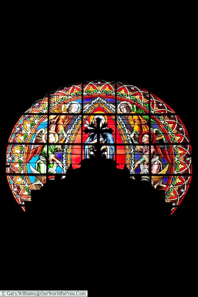 A vibrant, arched stained glass window from Sisteron in Provence, France, glows with rich reds, blues, greens, and yellows, depicting religious figures with halos in an ornate, traditional design. The intricate details and vivid colours of this historic French artwork are sharply contrasted against a deep black background, highlighting its beauty and craftsmanship.