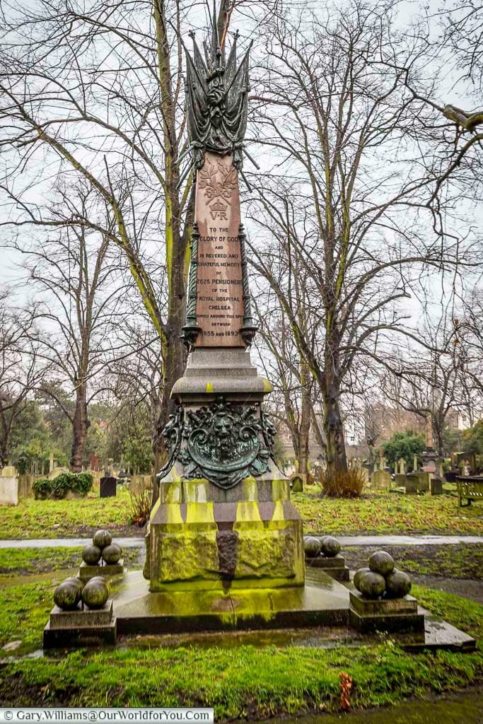 This image shows the Chelsea Pensioners Memorial in Brompton Cemetery, United Kingdom, a tall, ornate monument dedicated to the advanced class pensioners of the Royal Hospital Chelsea between 1855 and 1893. The memorial features intricate carvings, inscriptions, and is surrounded by cannonballs at its base, set against a backdrop of bare trees and other gravestones.