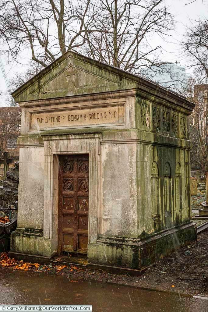 This image shows the weathered stone family tomb of Benjamin Golding M.D. in Brompton Cemetery, United Kingdom, on a rainy day. The mausoleum features a heavy, ornate wooden door and inscriptions on the stone facade, with bare trees visible against the overcast sky in the background.