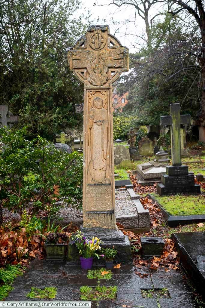 This image captures a detailed, weathered Celtic cross monument in Brompton Cemetery, United Kingdom, adorned with intricate carvings and standing on a stone base. A small purple pot of flowers rests at the foot of the cross, surrounded by fallen leaves and other gravestones in the background.
