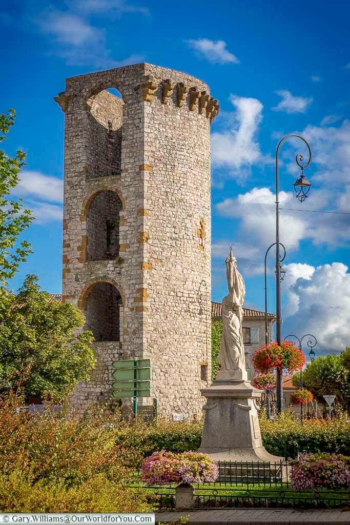 A tall, cylindrical stone tower, identified as the Médisance Tower in Sisteron, Provence, stands prominently with arched window openings and a crenellated top against a vivid blue sky with fluffy white clouds. In the foreground, a classical white statue on a plinth is surrounded by colourful flowerbeds and green bushes, with ornate street lamps visible to the right.