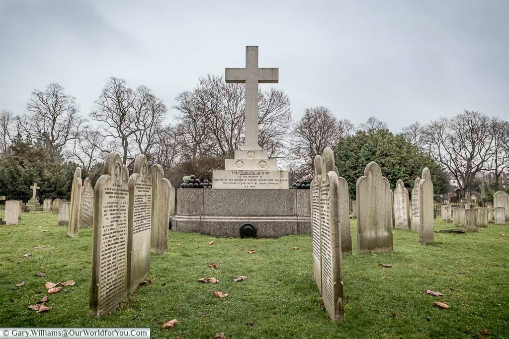 This wide shot shows a section of Brompton Cemetery in the United Kingdom under a cloudy sky, featuring a large stone cross monument surrounded by numerous smaller, weathered headstones on a grassy area with fallen leaves. Bare trees stand in the background, adding to the solemn atmosphere of the Victorian-era cemetery.