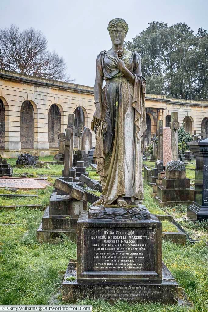 This eye-level shot shows the memorial statue of Blanche Roosevelt Macchetta in Brompton Cemetery, United Kingdom, on a wet day. The bronze statue depicts a woman holding a rose, standing atop a stone pedestal with inscriptions detailing her life.