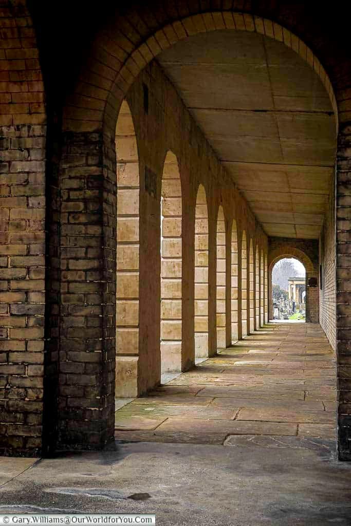 This image shows a long, covered walkway, likely a colonnade, at Brompton Cemetery in the United Kingdom. The walkway features a series of arched openings supported by brick and stone pillars, leading towards an open area with gravestones visible in the distance under a diffused light.