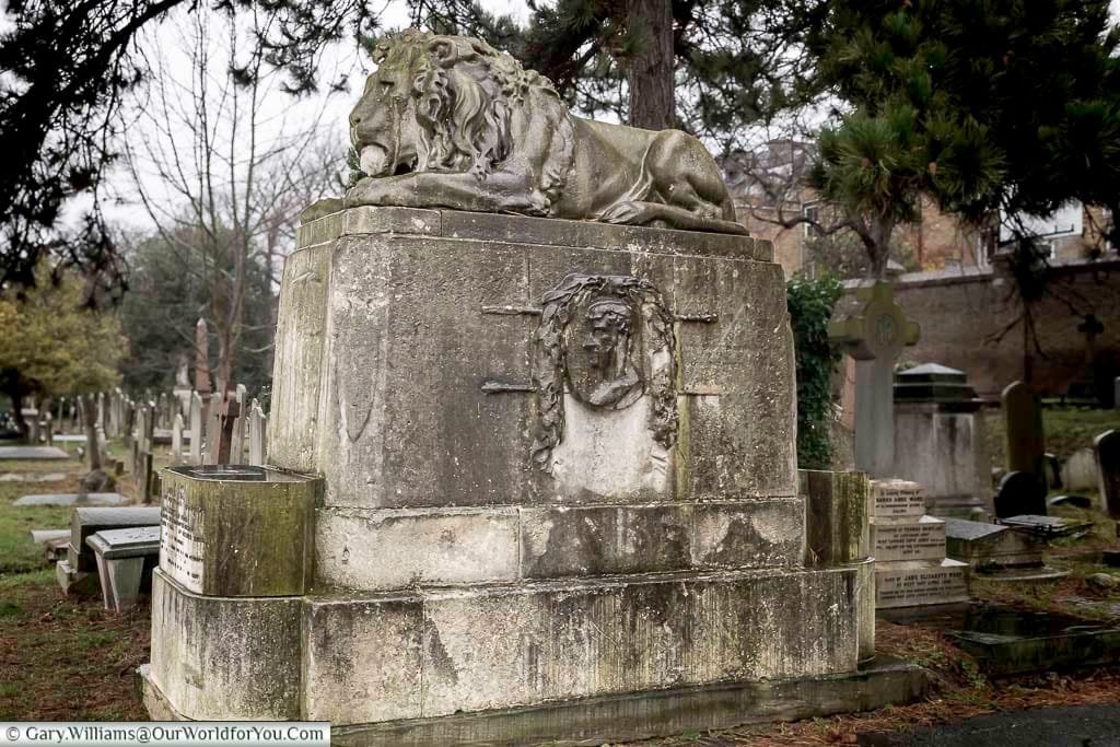 This close-up shot shows an ornate, weathered stone monument in Brompton Cemetery, United Kingdom, featuring a reclining lion sculpture on top. Below the lion, the monument is adorned with a carved face surrounded by foliage, and the base is composed of large, aged stone blocks.