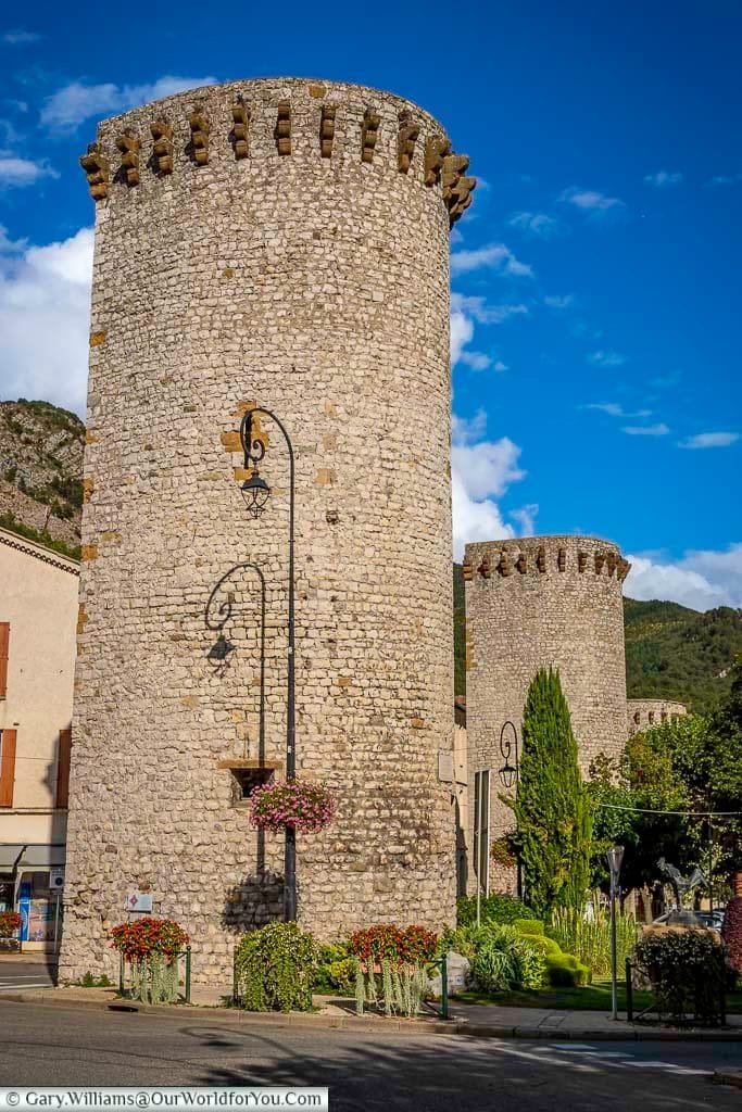 Three imposing medieval stone towers with crenellated tops stand prominently in Sisteron, Provence, France, showcasing the town's rich history and iconic architecture. Surrounded by flowering plants, classic street lamps, and glimpses of nearby buildings and distant hills, these historic French landmarks are set against a bright blue sky with fluffy white clouds.