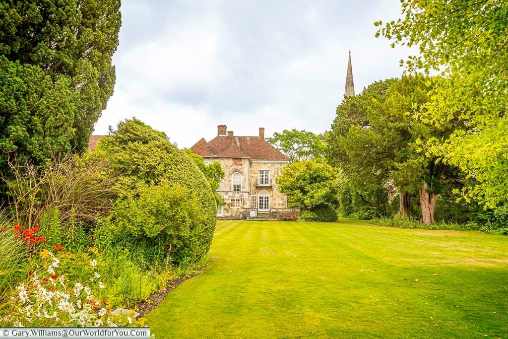 A lush green lawn extends towards the historic Arundells house, a multi-storey building with prominent chimneys and light-coloured stone, nestled amongst mature trees. In the foreground, a vibrant flowerbed blooms with red and white flowers, while the spire of Salisbury Cathedral is just visible above the trees in the distance, all under an overcast sky.