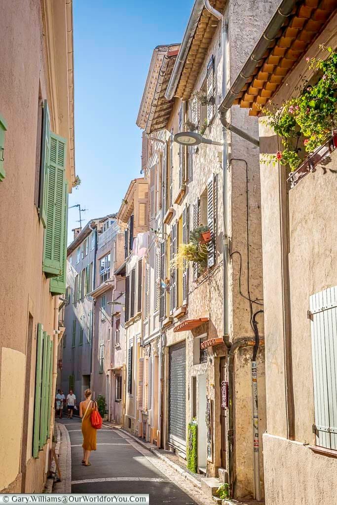 A colorful street in Antibes, France, features pastel-hued buildings with classic shutters, flower-filled balconies, and a pedestrian in a yellow dress walking through the quaint scene. The vibrant architecture and serene atmosphere highlight the Mediterranean charm of this historic town.