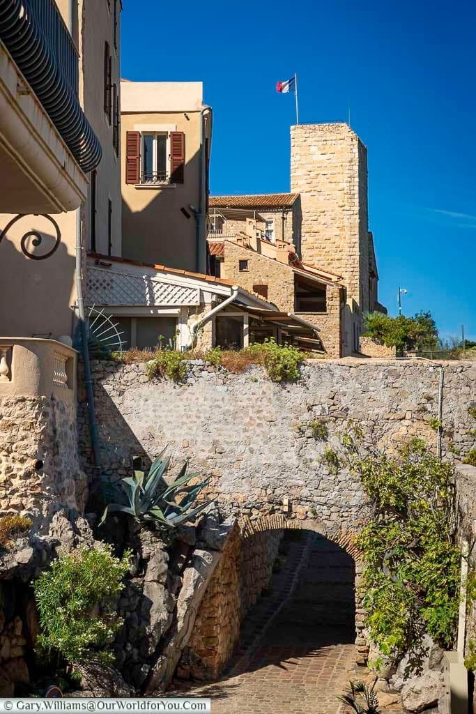 A picturesque street scene in Antibes, France, featuring weathered stone buildings, a narrow archway, and the French flag waving atop a historic tower. The composition highlights the town's architectural charm and timeless ambiance under a bright Mediterranean sky.