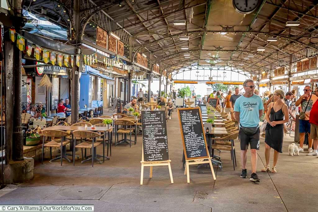 A bustling open-air market scene in Antibes, France, showcases diners enjoying Mediterranean dishes like mussels and fries, Caesar salad, and beef tartare beneath a rustic covered structure. Chalkboard menus and vibrant activity create a lively, authentic atmosphere ideal for lifestyle, travel, and culinary content.