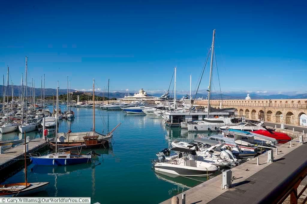 The marina in Antibes, France, features rows of sleek yachts and sailboats docked along the stone piers under a vibrant blue sky. This coastal scene captures the elegance and charm of the French Riviera, perfect for travel and lifestyle content.