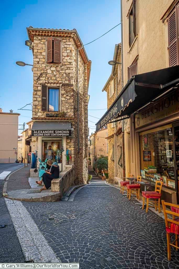 A charming street view in Antibes, France, featuring the Alexandre Simcini Galerie d'Art Contemporain and BeerShop06 set against traditional stone architecture. A seated figure near the gallery entrance adds a touch of everyday life to this picturesque and culturally rich scene.