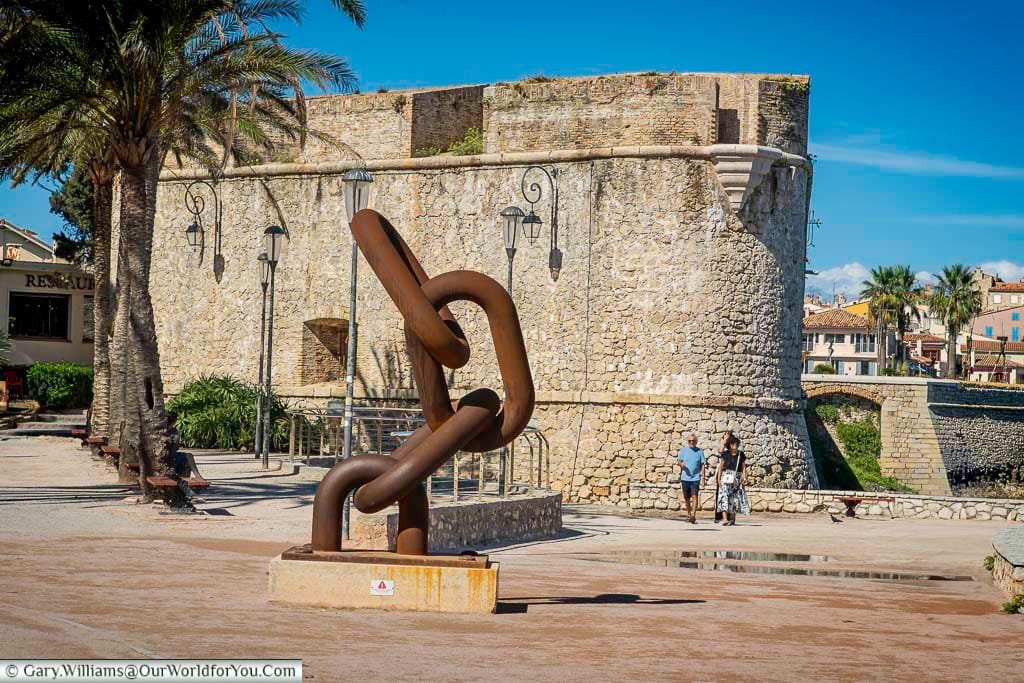 A striking sculpture of interlocking metal chains stands in front of the historic Bastion Saint-André in Antibes, France, blending modern art with ancient stone fortifications. Palm trees, cobbled walkways, and Mediterranean architecture complete the scene, perfect for showcasing Antibes’ blend of culture, history, and coastal charm.