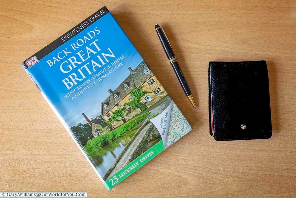 A travel planning flat lay featuring the DK Eyewitness "Back Roads Great Britain" guidebook, a pen, and a black wallet on a wooden surface. Perfect for organizing scenic road trips across the UK, this book offers 25 leisurely drives with charming hotels, authentic cuisine, and detailed pull-out maps.