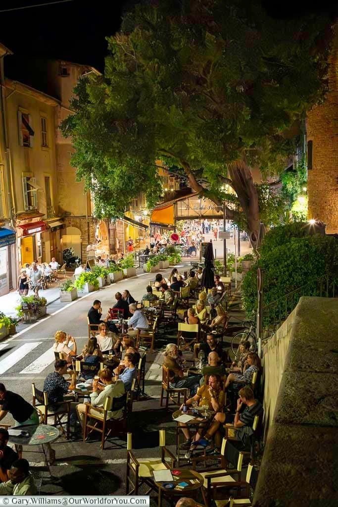 An atmospheric evening café scene in Antibes, France, where locals and visitors enjoy outdoor dining under warm streetlights and leafy green surroundings. The image captures the lively charm of Mediterranean nightlife, highlighting the town's inviting ambiance and social culture.