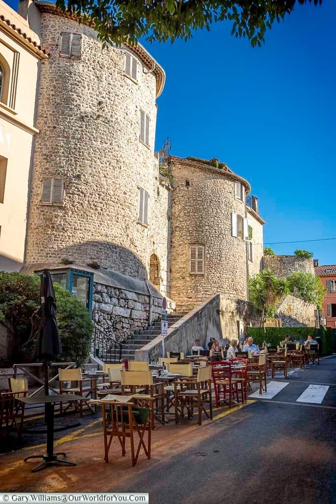 A quaint outdoor café in Antibes, France, features tables and chairs set along a narrow cobblestone street beside centuries-old stone buildings. The inviting scene captures the town’s relaxed Mediterranean vibe, enhanced by lush greenery and a bright blue sky.