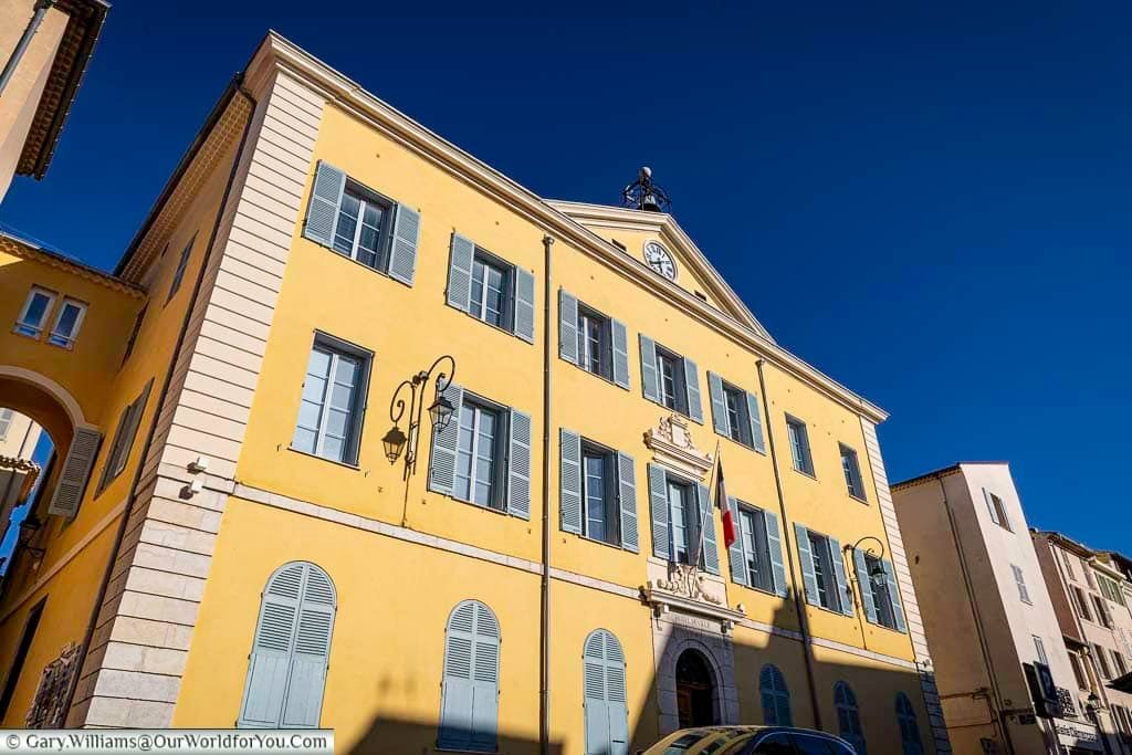 A striking yellow building with vivid blue shutters and a central clock tower stands prominently in Antibes, France, beneath a clear Mediterranean sky. This architectural landmark, adorned with a French flag and intricate detailing, showcases the colorful charm of the Côte d’Azur.