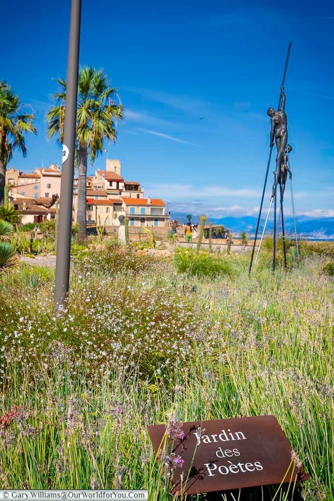 A vibrant view of Jardin des Poètes in Antibes, France, featuring wildflowers, palm trees, and a striking modern sculpture set against the old town's terracotta rooftops and the distant Alps. This peaceful garden by the Mediterranean Sea is a hidden gem for visitors exploring Antibes and the French Riviera.