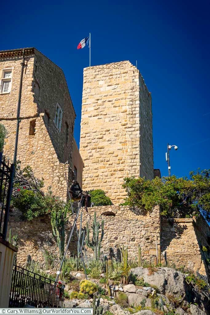 A historic tower and stone building in Antibes, France, rise above a cactus-filled garden featuring a modern sculpture of a figure perched on tall stilts. The scene blends artistic creativity with architectural heritage under a vivid blue Mediterranean sky.