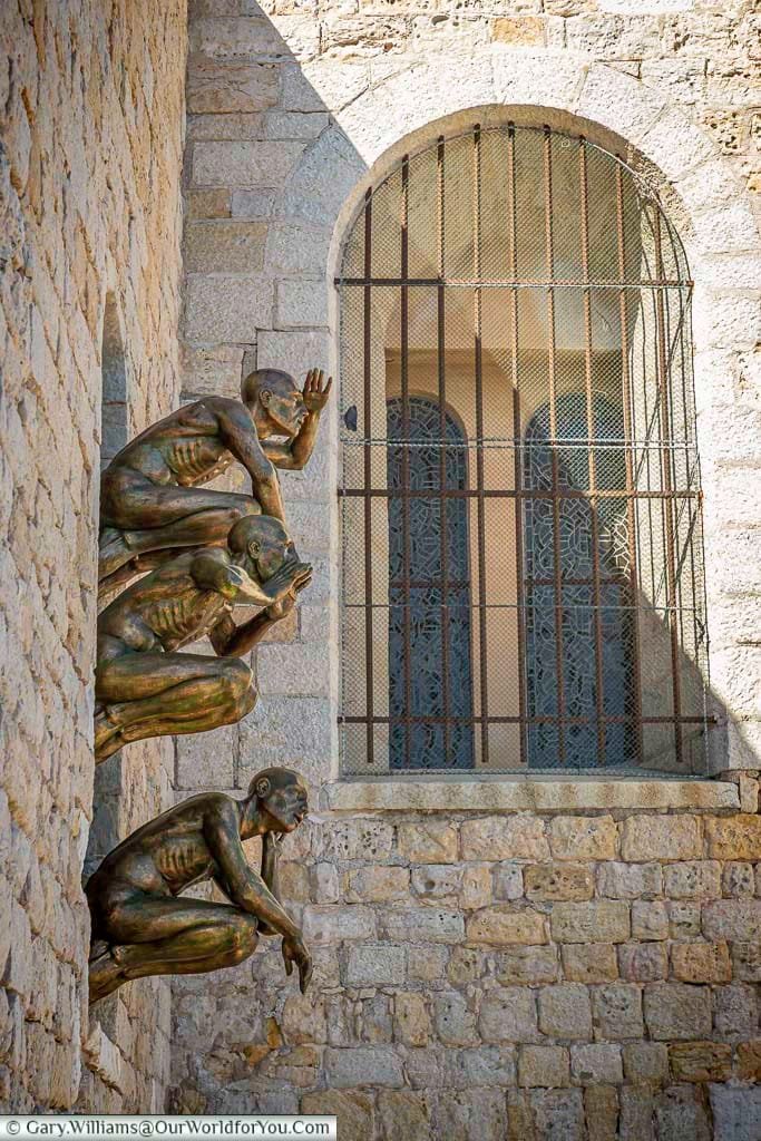 Four bronze crouching figures are mounted on the stone wall of the Musée Picasso in Antibes, France, gazing through a barred window. This artistic installation offers a striking juxtaposition of contemporary sculpture with the historic architecture of the coastal museum.