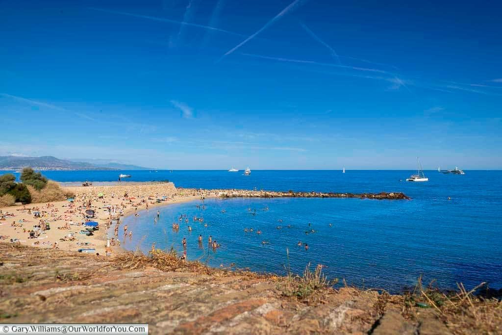 A vibrant beach scene in Antibes, France, showcasing sunbathers along the sandy shore and swimmers enjoying the turquoise Mediterranean waters. The image captures the lively coastal atmosphere with boats in the distance and clear skies overhead, ideal for travel and leisure content.