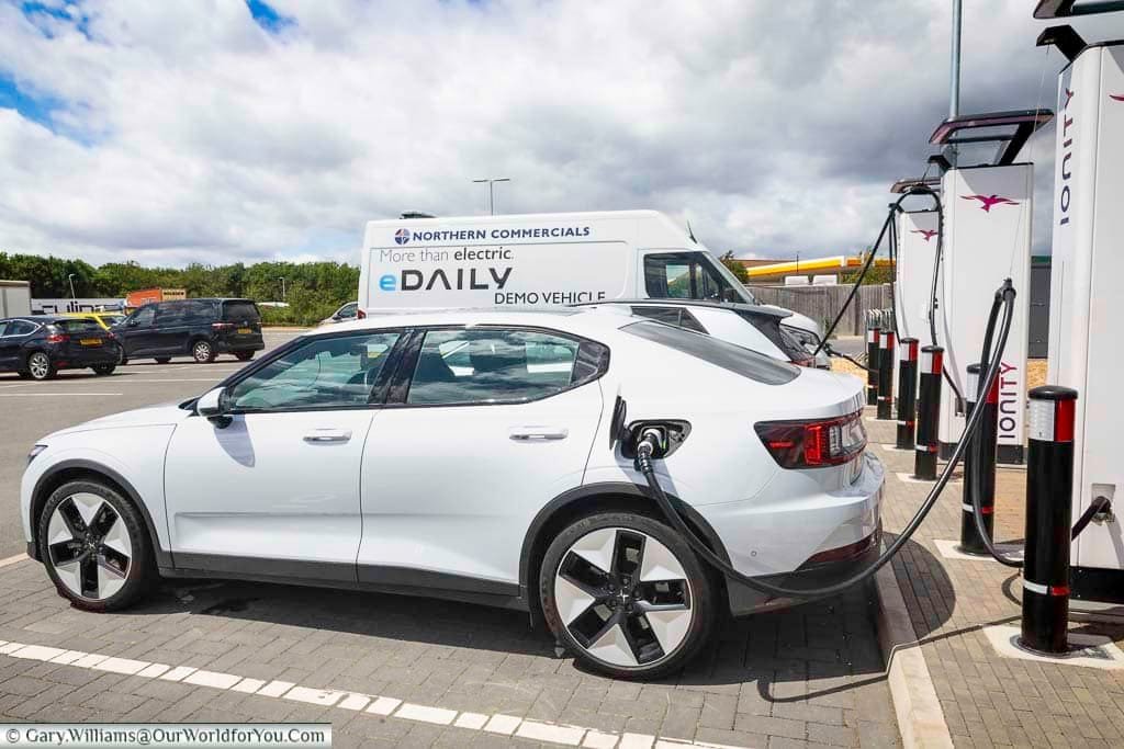 A white Polestar 2 electric vehicle is seen charging at IONITY rapid chargers at Cambridge Services, showcasing the growing UK EV infrastructure. This convenient motorway stop highlights the ease and efficiency of sustainable travel during an electric vehicle road trip across England.