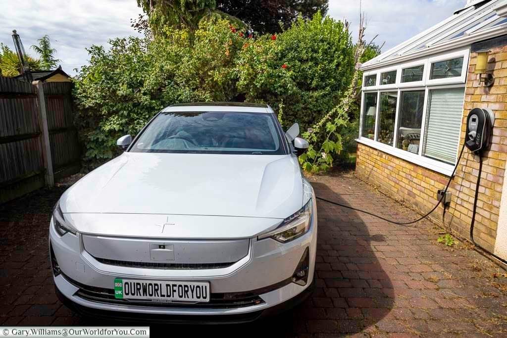 A white Polestar 2 electric vehicle is parked on a brick driveway, charging via a home EV wall charger mounted on a conservatory wall. This image highlights the convenience of home charging for electric vehicle owners and promotes sustainable living and road trip readiness in the UK.