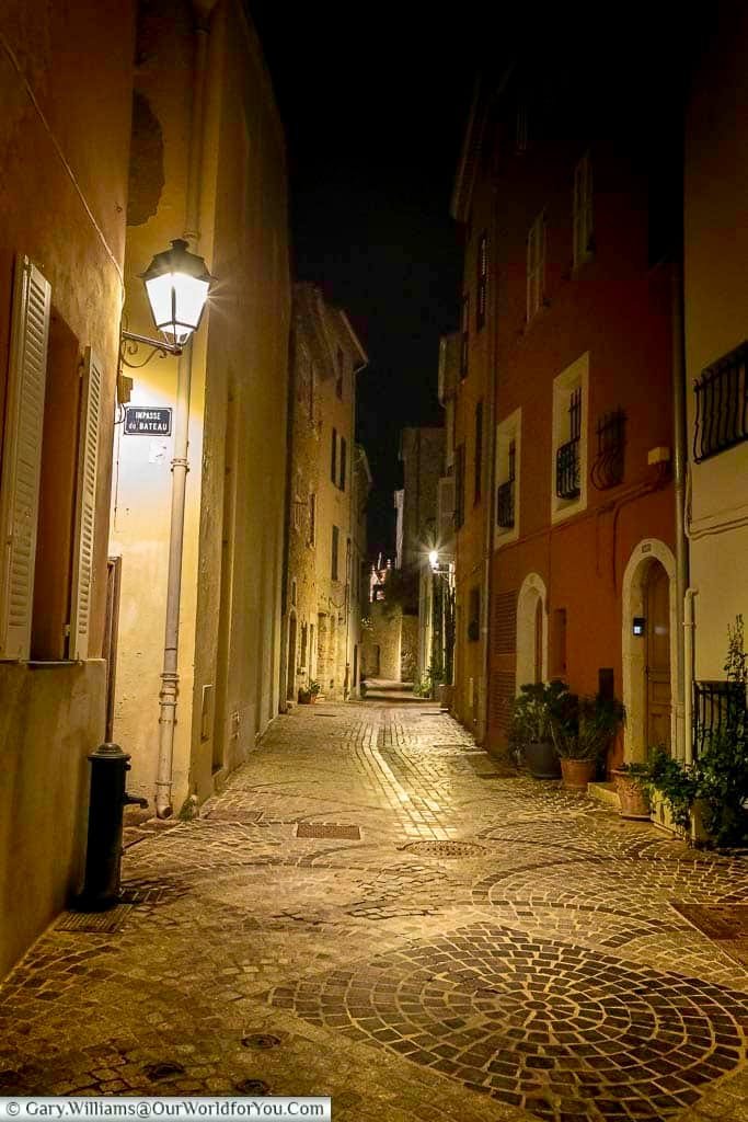 A picturesque nighttime alley in Antibes, France, showcases cobblestone pavement, softly lit streetlamps, and historic buildings adorned with shuttered windows and potted greenery. This inviting scene reflects the romantic charm and timeless beauty of the town’s Mediterranean architecture.