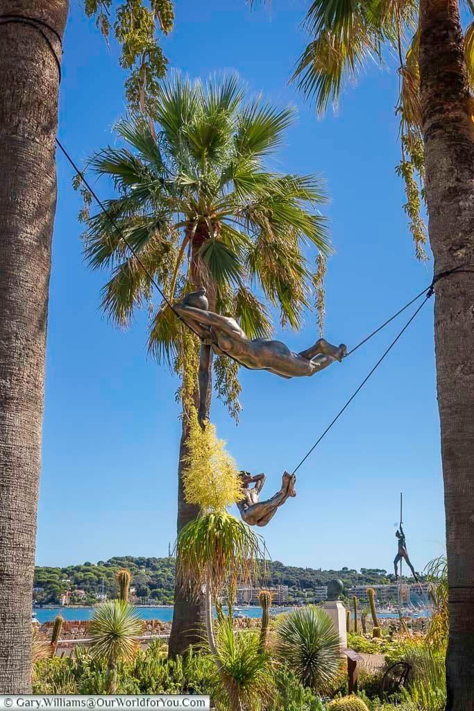 Sculpture installation in Antibes, France, showcasing dynamic bronze acrobats suspended between palm trees under a vivid blue sky. The artwork enhances the scenic coastal view, with lush greenery and the Mediterranean Sea in the background.