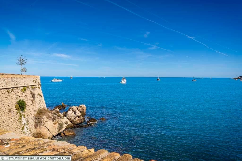 A scenic view of the Mediterranean Sea from Fort Carré in Antibes, France, featuring yachts and sailboats anchored in crystal-clear blue waters. The historic stone fort and rugged shoreline frame the tranquil coastal landscape under a bright, cloudless sky.