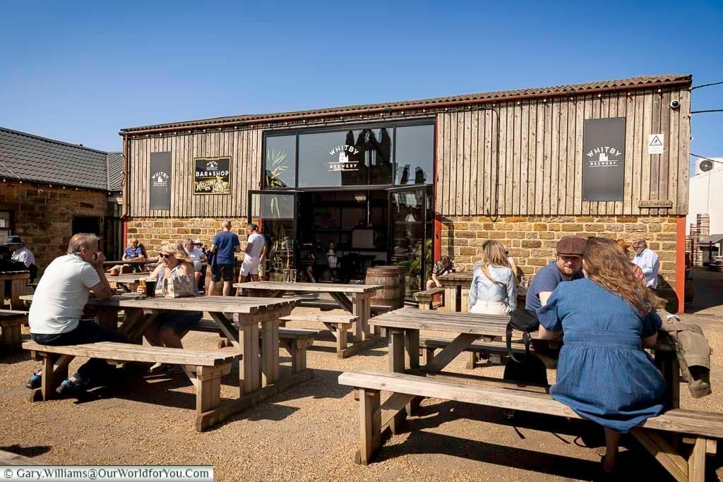 Visitors enjoying drinks at outdoor wooden tables outside Whitby Brewery in Yorkshire, England, with stone architecture, signage, and a clear blue sky framing the vibrant social scene at this popular coastal craft beer destination.