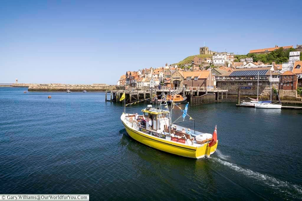 A vibrant yellow tour boat cruises through the picturesque Whitby Harbour in Yorkshire, with charming red-roofed cottages and the historic St Mary's Church overlooking the scene. This iconic coastal town offers rich maritime heritage, scenic views, and a perfect stop on any Yorkshire road trip.