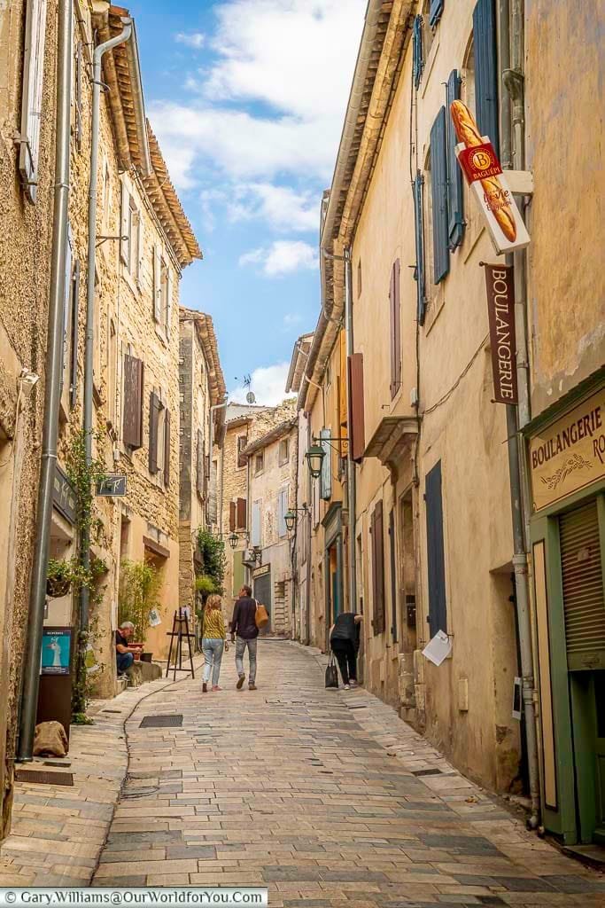 Quaint cobbled street in Ménerbes, Provence, lined with rustic stone houses, artisan shops, and a traditional boulangerie. This charming village scene perfectly captures the relaxed pace and authentic beauty of southern France.