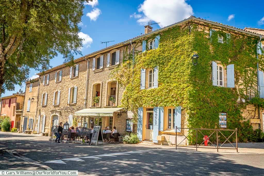 A charming village square in Goult, Provence, features ivy-covered stone buildings, pastel shutters, and a cosy café terrace buzzing with lunchtime diners. Bathed in golden sunlight, this idyllic scene captures the timeless allure of southern France and its relaxed, scenic way of life.