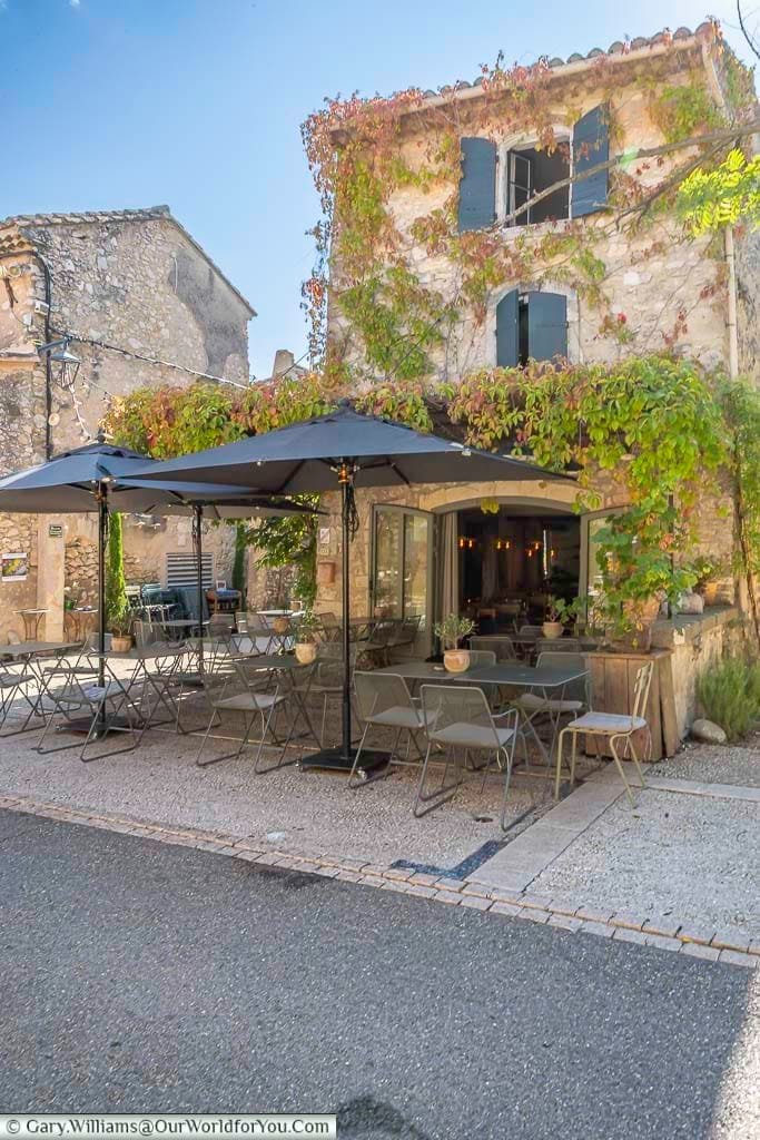 Charming café terrace in Eygalières, Provence, set against a rustic stone house draped in vibrant climbing vines. This peaceful village scene embodies the relaxed elegance and hidden beauty of southern France.